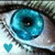 A close-up of a blue eye with visible eyelashes and skin texture, featuring a small teal heart symbol in the lower-left corner.