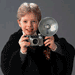 A person in a black coat holds a silver camera with a flash, smiling while text overlays change across frames.