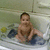 A child sits in a bathtub with blue water, holding a yellow object and dark clothing item while looking forward.