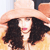 A pixelated close-up of a person wearing a light-colored wide-brimmed hat, dark hair, and a pink lip color, with a blurred background.