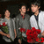 Three pixelated figures stand side-by-side holding red flowers, with the left figure in gray shirt and green pants, middle in dark jacket over red collar, right in white coat with black tie.
