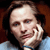 Pixelated close-up of a person with long dark hair resting their head on a hand, wearing a light blue shirt against a brown background.