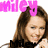 A pixelated image of a woman with brown hair, smiling and holding her hand near her face, overlaid with pink text reading 'miley' at the top left.