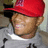 Pixelated close-up of a person wearing a red cap and light-colored shirt with dark text, set against a blurred background.