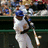 Pixelated image of a baseball player in white uniform swinging a bat, background shows blurred spectators and green stadium railings.
