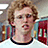 A pixelated image of a person with curly light brown hair wearing a white shirt with black collar, set against a bright pink background with blue text.