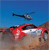 Red car with white accents on a brown terrain under a blue sky, with a helicopter in the background.