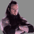 Pixelated static image of a person with long dark hair wearing a black top, seated against a gray background.