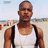 A person in a white tank top with black straps stands on a beach, the right side of their chest displays five pink pixelated shapes.