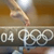 A static image shows a hand holding a silver torch above the Olympic rings and '04' text on a brown background.
