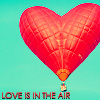 A red heart-shaped hot air balloon with a basket hangs against a light blue background, with text below reading LOVE IS IN THE AIR.