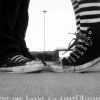 Black-and-white image of two people's lower legs and feet standing on a paved surface, one wearing striped pants and the other dark pants, with a blurred background featuring a tall pole.