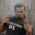 Pixelated static image of a person with blue hair and glasses wearing a dark shirt, making a hand gesture against a gray background.