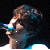 Pixelated static image of a person with brown hair and blue highlights against black background, wearing dark clothing with white accents.