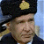 A pixelated close-up of a person wearing a dark hat with a gold emblem, light-colored hair, and a dark jacket against a light blue background.