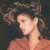 Pixelated image of a person with dark hair styled in an updo, wearing a light brown top against a dark background.