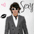 A person in a gray suit with sunglasses and brown hair stands against a white background, next to pink heart and black lip icon symbols.