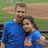 Two people in blue shirts pose closely together, one with an arm around the other, against a green and brown background.