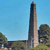 Tall, narrow, dark gray obelisk-like structure with light brown base against clear blue sky, surrounded by green trees and grassy terrain.