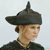 Pixelated image of a person wearing a dark conical hat with a small protrusion, light brown hair, and a dark collared garment against a light gray background.