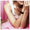 A person in a white sleeveless top and pink skirt rests their chin on a hand, wearing a beaded necklace and rings, with a pink curtain background.