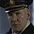 Static pixelated image of a person wearing a dark military-style cap with a gold emblem, a white collar, and a dark jacket against a muted blue background.