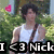 Pixelated image of a person in a white tank top with dark hair, standing outdoors near greenery and holding a pink heart-shaped object.