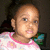 Pixelated image of a child with light brown skin wearing a white shirt with pink trim and green accents, set against dark background.