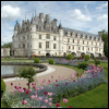 A static image of a large white chateau with multiple towers and windows, surrounded by a landscaped garden with pink flowers, a curved pathway, and a pond under a partly cloudy sky.