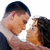 Two people in close physical proximity, one with short dark hair and the other with long dark hair, facing each other against a white background.