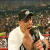A pixelated static image shows a person with dark hair wearing a white top, holding an object near their face against a blurred background.