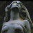 Pixelated static image of a figure with long flowing hair, wearing a hooded garment and seated in profile against dark background.