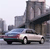 Pixelated image of a light-colored sedan with dark accents driving past a large stone bridge structure under a pale sky.