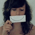 A person with dark wavy hair holds a white envelope-shaped object in front of their mouth, wearing a sleeveless top against a light background.
