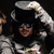 A person in a black top hat and dark clothing holds the hat with one hand while holding an object, surrounded by text elements on a black background.