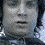 A pixelated animated sequence showing a person in a dark jacket with a light-colored collar, holding a small object near their chest, with background elements shifting from blue to gray tones.