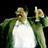 Pixelated static image of a person in green jacket and white shirt holding an object against black background.