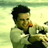 Pixelated image of a person with dark hair wearing a light green jacket over a white shirt, holding an object near their mouth against a pale yellow-green background.