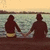 Two silhouetted figures holding hands on a wooden pier at sunset, facing water with a gradient sky.