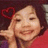 A person with dark hair and a red top, smiling while resting their hand on their cheek, with a small red heart symbol above the head.
