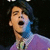 Pixelated image of a person with dark hair wearing a purple garment, mouth open in an expressive pose against a black background.