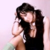A person with long dark hair wearing a black sleeveless top and light green socks sits with one knee raised, hand resting on head, against a plain light background.