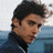 Pixelated portrait of a person with dark hair wearing a dark jacket over light blue clothing against a blurred background.