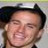Pixelated close-up of a person's face with dark hair, closed eyes, and a broad smile showing teeth, set against a light background.