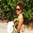 A pixelated image shows a person with brown hair tied up, wearing sunglasses and a sleeveless top, standing outdoors near green foliage.