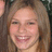 A pixelated close-up of a smiling person with light brown hair, visible teeth, and rosy cheeks against a dark background.