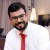 Pixelated image of a person in white shirt and red tie with dark hair, beard, and glasses against blurred background.