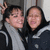 Two people pose closely together, one making a peace sign with their right hand while both wear dark jackets over light-colored shirts against an indistinct background.