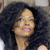 A pixelated close-up of a person with dark curly hair, light skin, and a neutral expression, wearing a dark top against a blurred background.