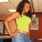 A person in a yellow crop top and blue jeans stands with one hand on their hip, holding a bottle near a wooden cabinet.